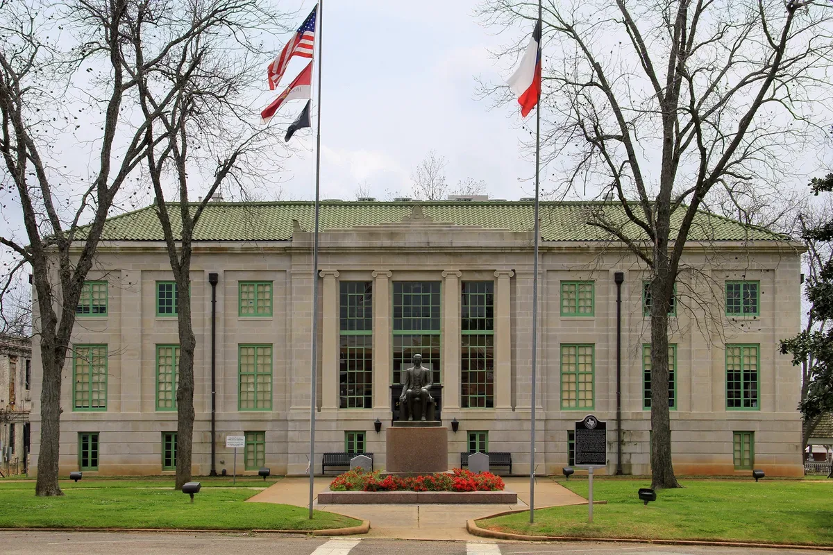 The San Augustine County Courthouse, a two-story pale stone building with a green tile roof and tall green-framed windows, behind a lawn with flagpoles, bare trees, a seated bronze statue on a pedestal and a historical marker