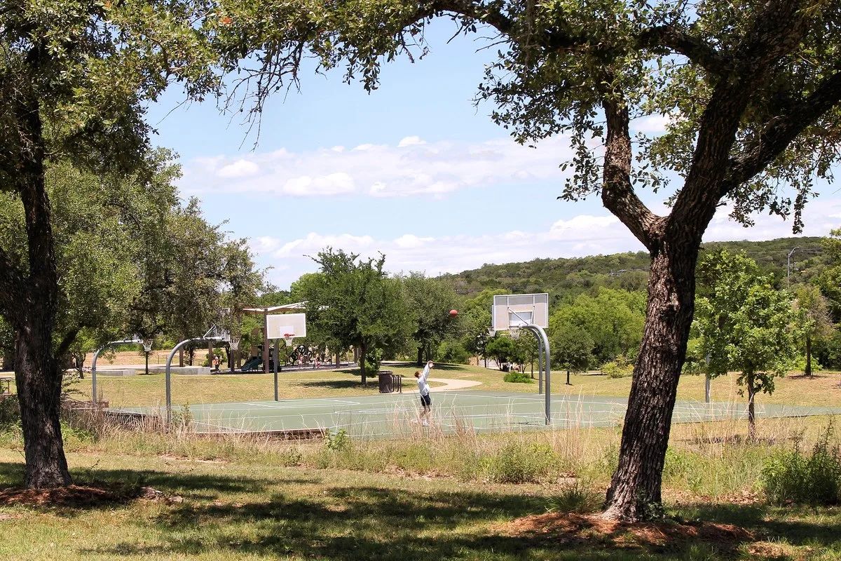 Basketball courts and a playground under live oak trees at Bee Cave Central Park, with green hills behind