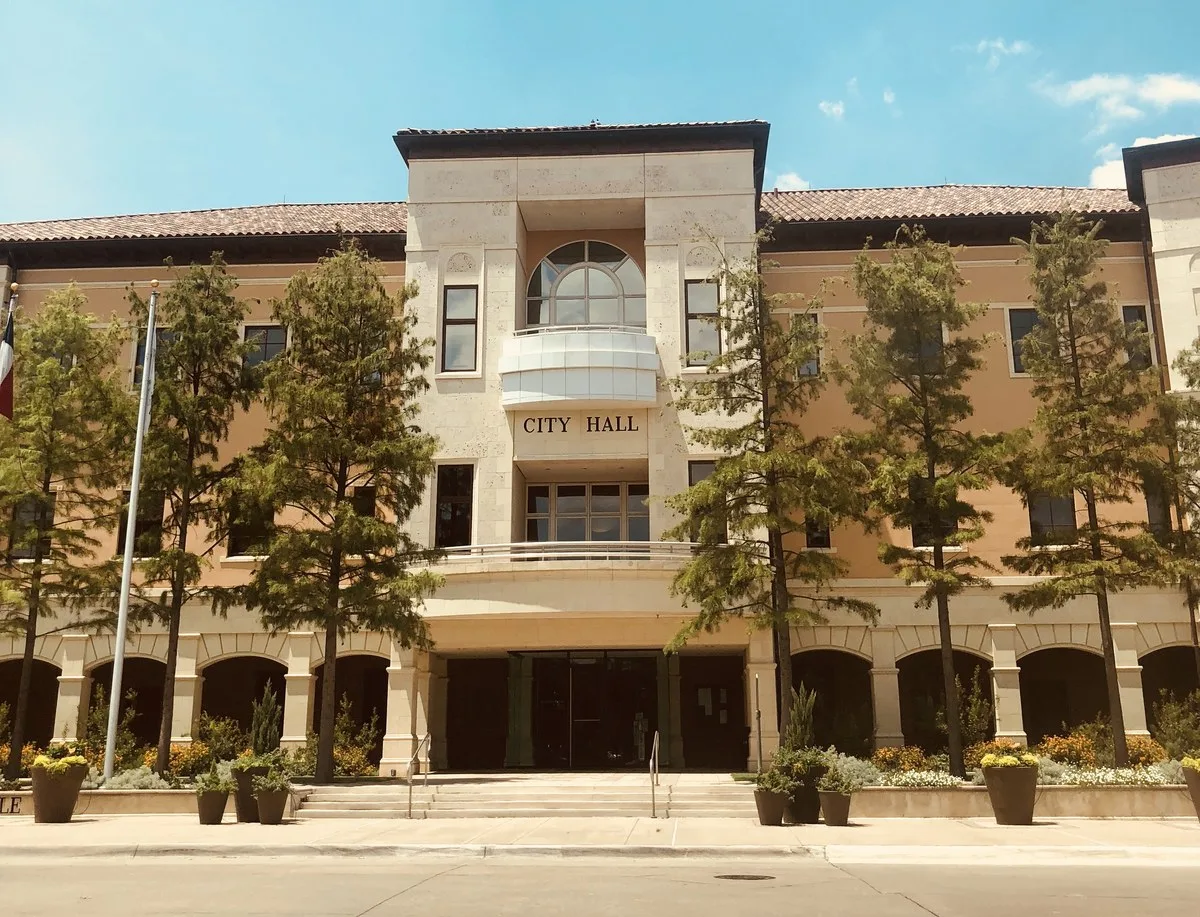 A tan stone and stucco city hall building with an arched window, a flagpole and young trees out front