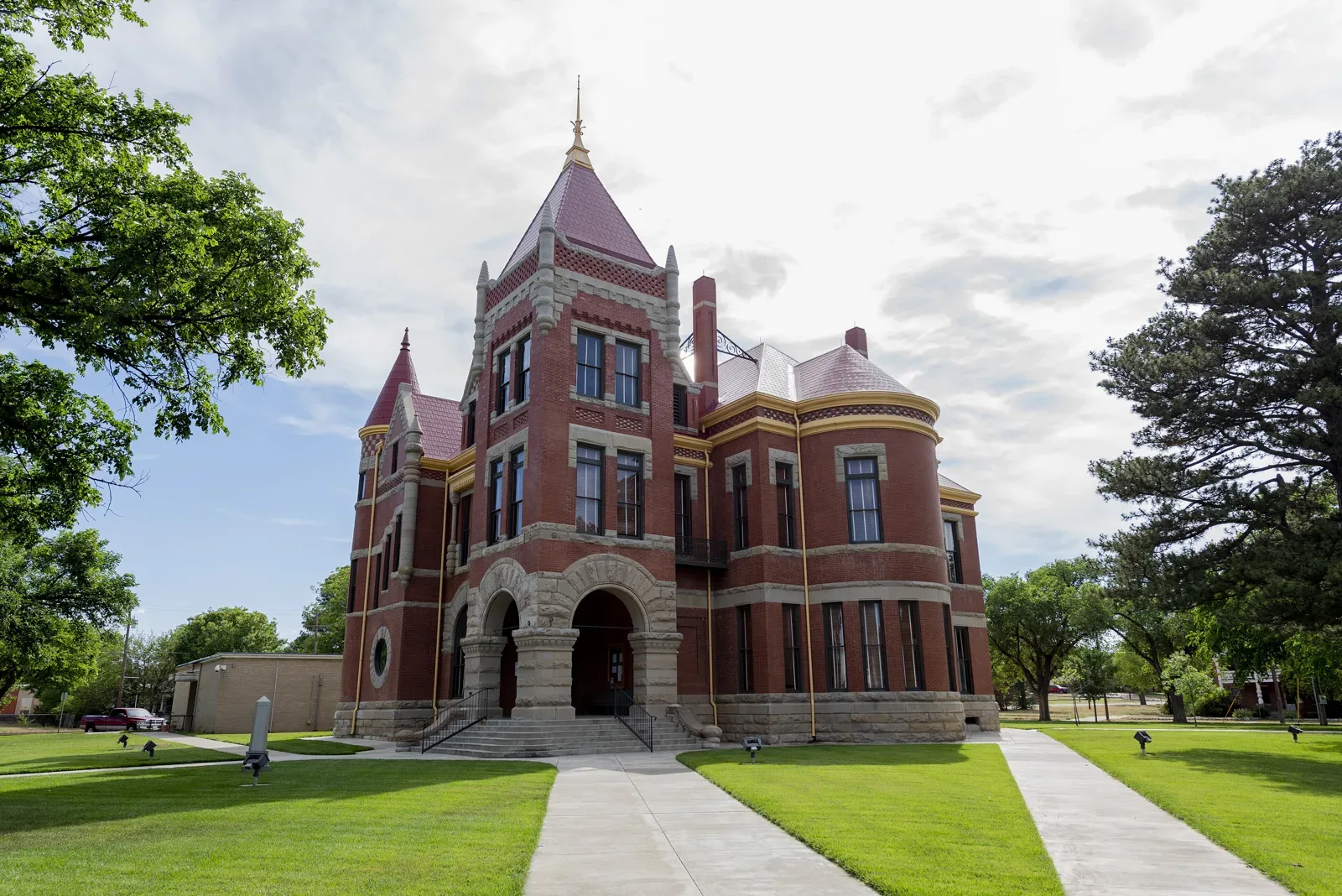 The Donley County Courthouse in Clarendon, Texas, a red brick Romanesque Revival building with limestone trim, a tall pointed central tower, a smaller corner turret with a conical roof, and a rounded arched entrance, behind a green lawn and sidewalks under a partly cloudy sky