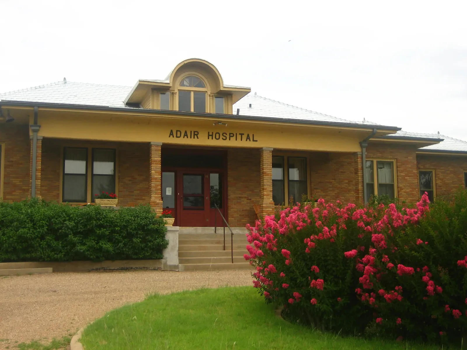 A one-story tan brick building with a hipped white metal roof, an arched dormer window and a deep front porch with brick columns and a sign reading Adair Hospital, with red front doors at the top of concrete steps and a pink crape myrtle in bloom in the foreground