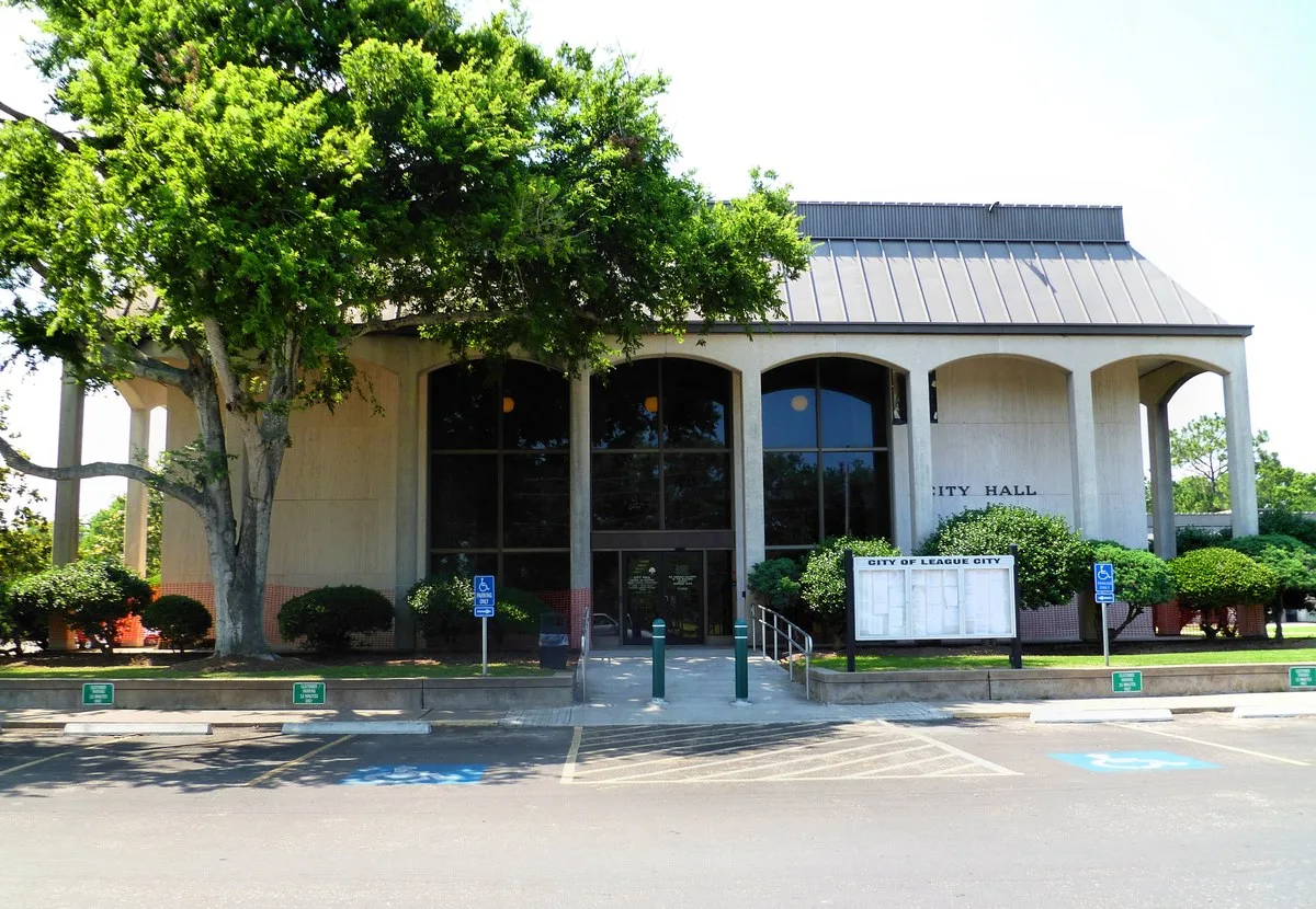 League City City Hall, a pale concrete building with tall arched windows behind a large shade tree