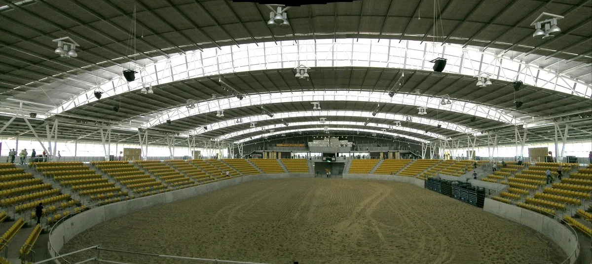 A large covered arena with a dirt floor, curved steel roof trusses and yellow stadium seats on both sides