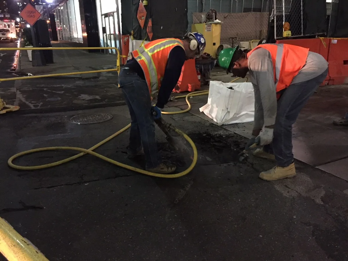 Two workers in orange safety vests and hard hats break up pavement on a city street at night