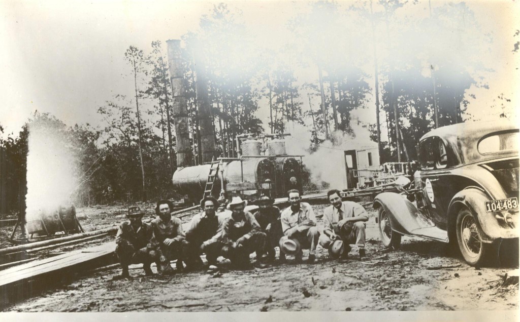 Oil-field crew crouched in front of boilers and smokestacks in the East Texas pines, with a 1934 Texas license plate visible on a parked car