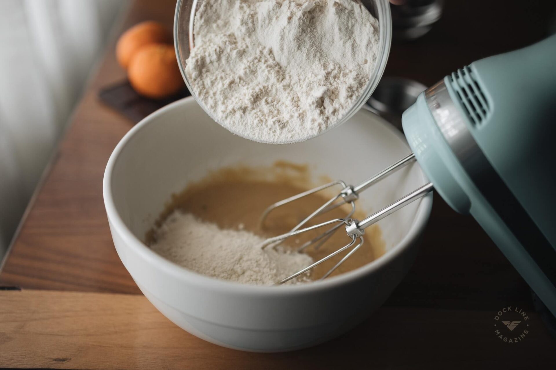 Flour being poured into a white mixing bowl beside a hand mixer, with oranges visible in the background