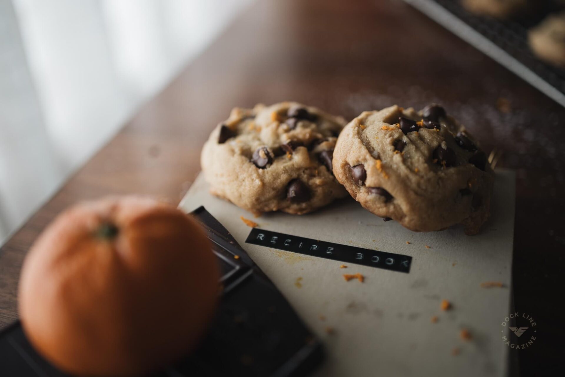 Two finished orange chocolate chip cookies on a recipe book beside a whole orange and dark chocolate bar