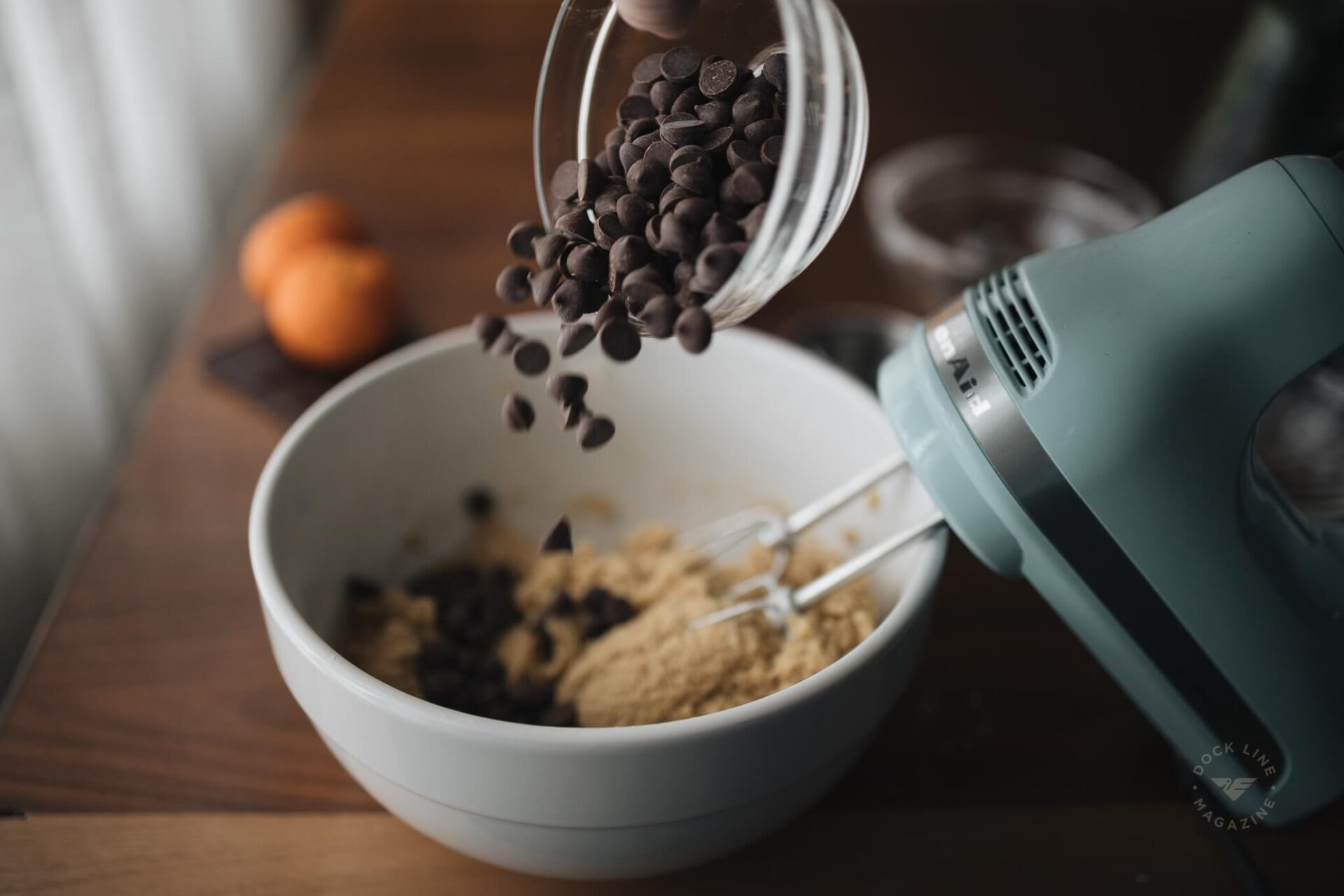 Dark chocolate chips cascading from a glass container into the cookie dough in a white bowl